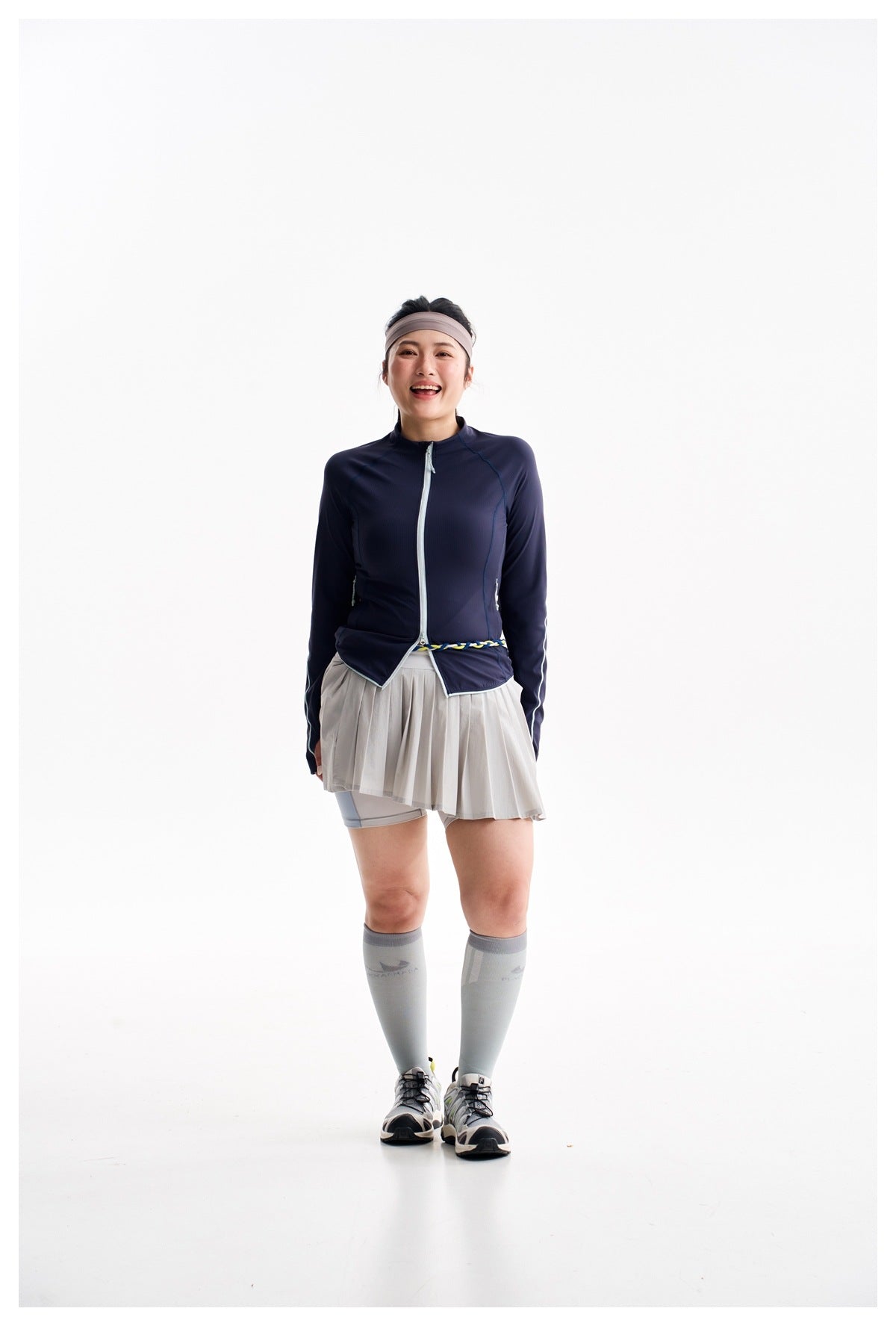 Model wearing a navy zip-up athletic jacket, gray pleated skort, gray knee-high socks, and running shoes.