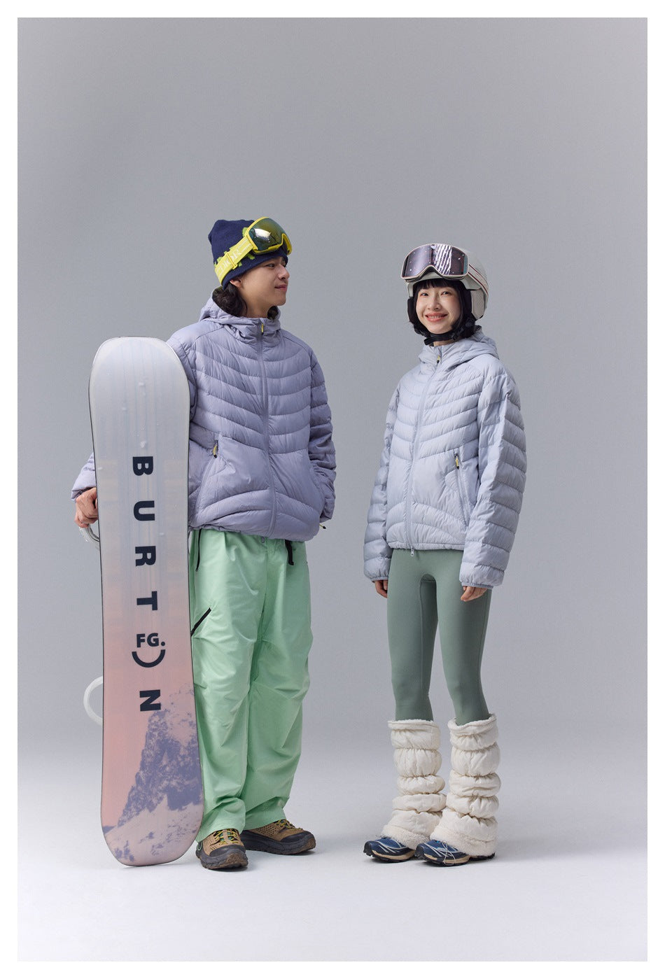 Two models wear pale lavender puffer jackets with snow gear beside a snowboard in a studio.