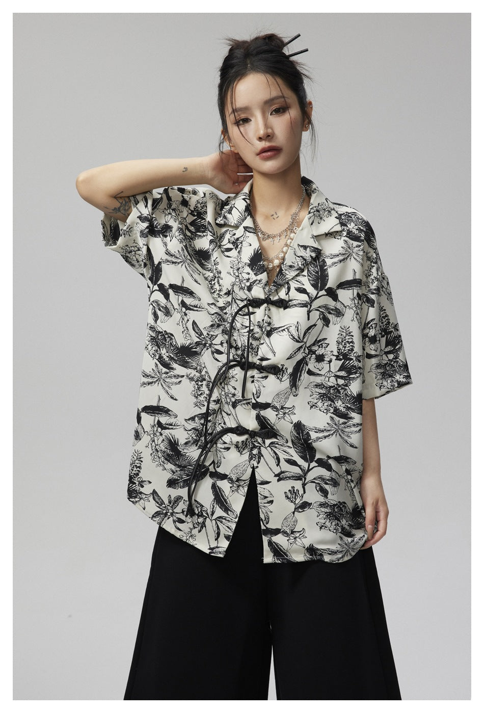 Model wearing an ivory short-sleeve shirt with black botanical print, black knotted closures, and wide black trousers.