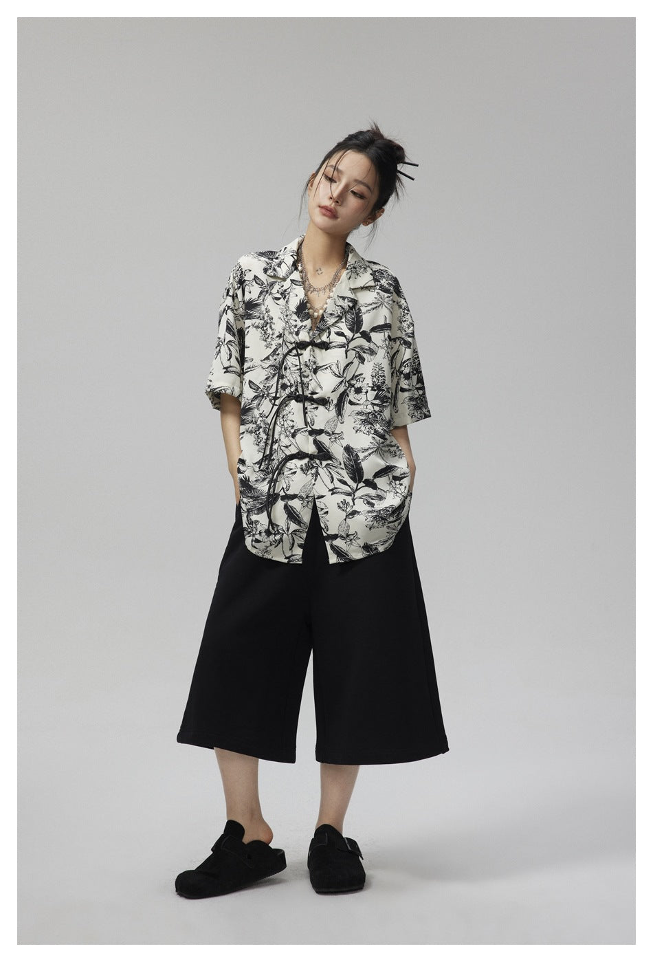 Model wearing an ivory botanical-print shirt with black frog closures and black wide-leg cropped pants.