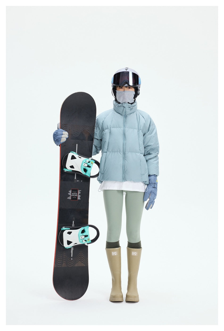 Front view of a person in a pale blue puffer jacket, sage leggings, gloves, and ski gear beside a snowboard.
