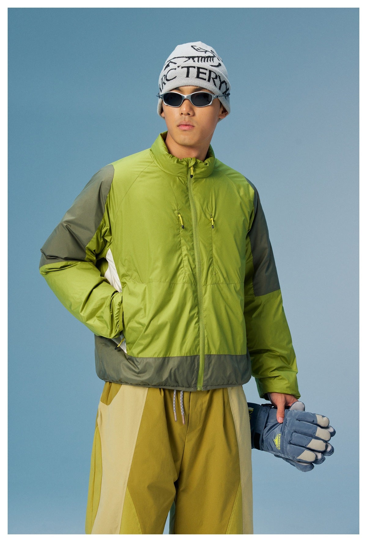 Male model wearing a lime green zip jacket with gray sleeve and hem panels, white side inserts, and mustard trousers.