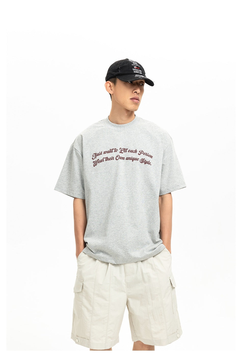 Model wearing a light gray T-shirt with burgundy script, cream cargo shorts, and a black cap.