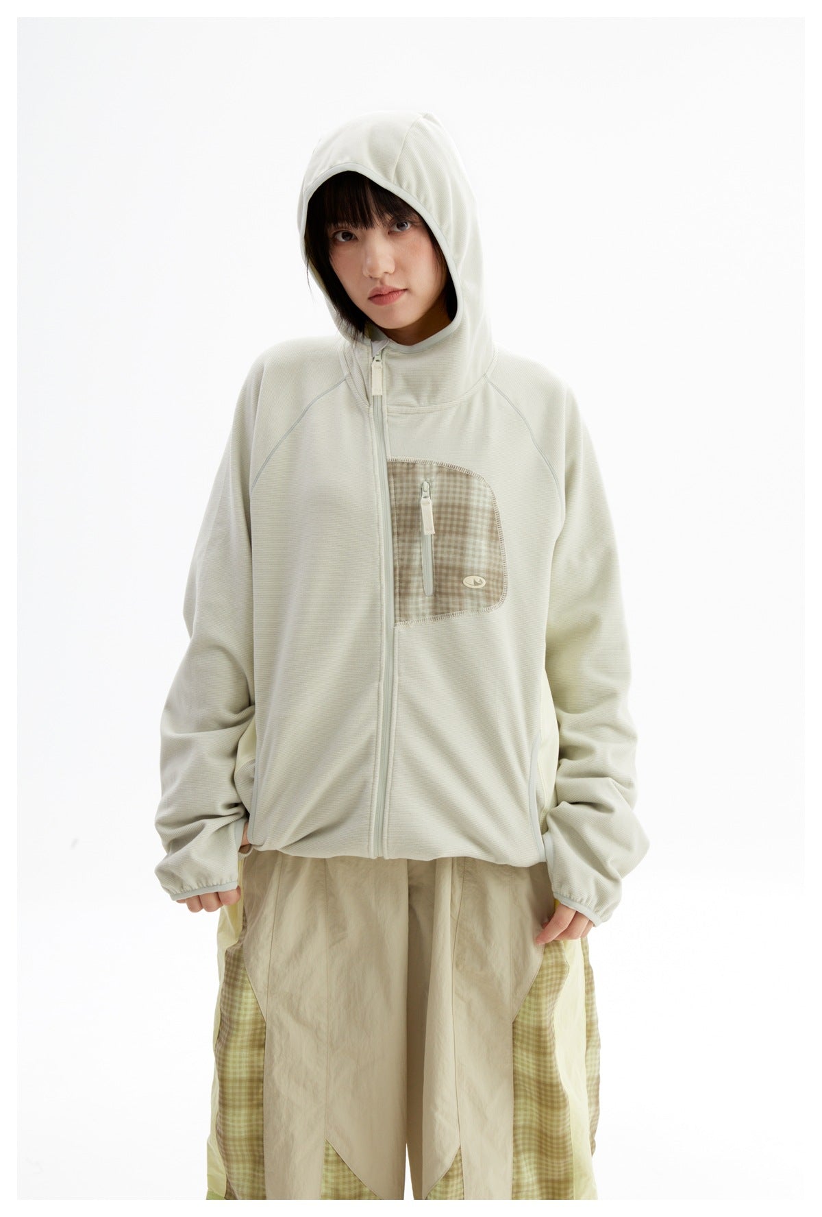 Model wearing an ivory hooded zip jacket with a checked chest pocket and oversized beige-and-green checked trousers.