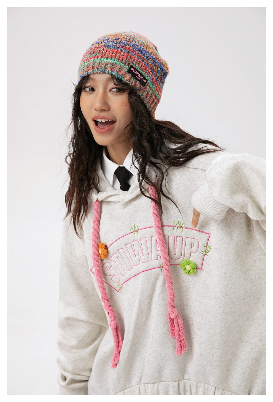 Close view of a model wearing a light gray hoodie with pink drawstrings and floral lettering and a colorful knit beanie.