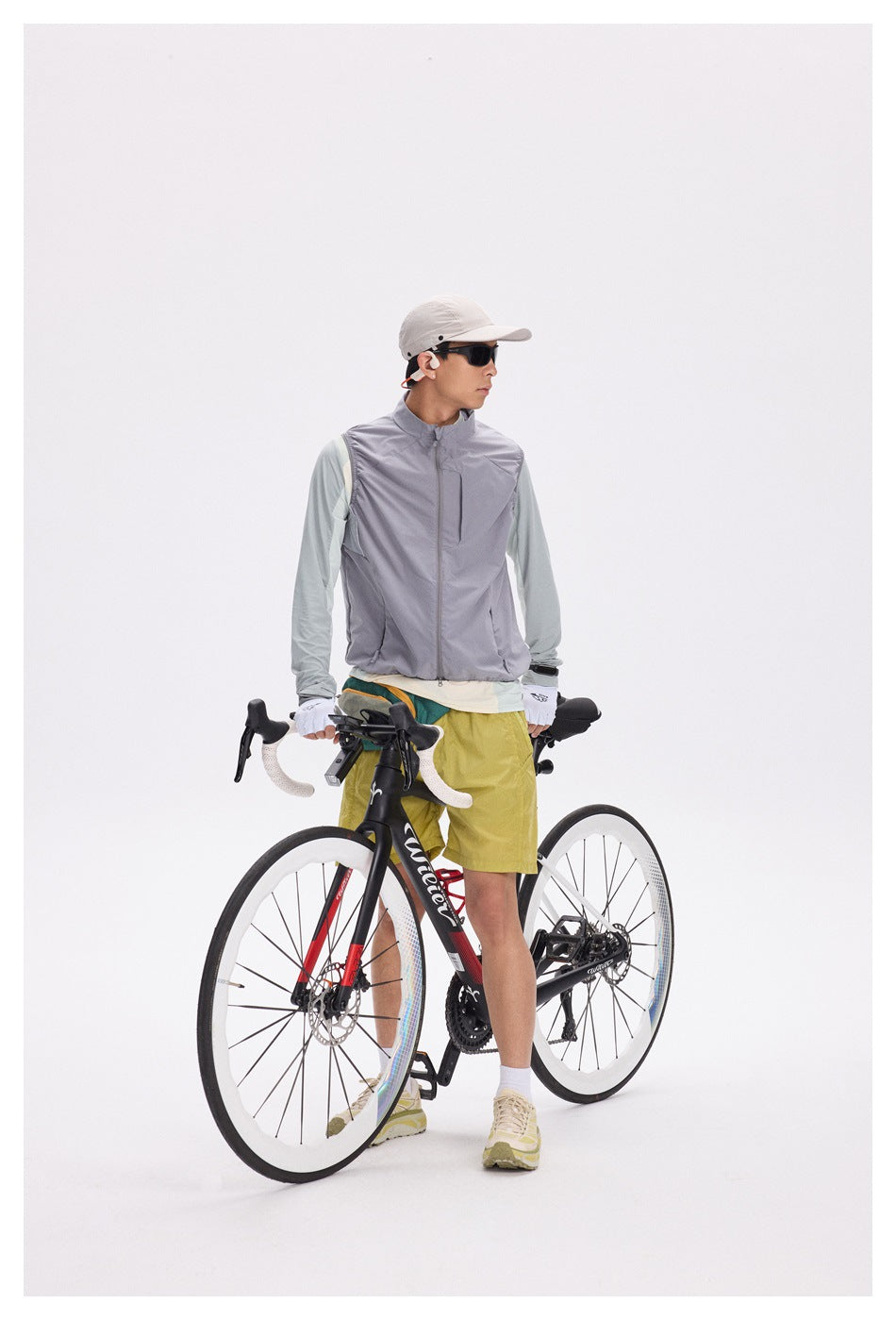 Cyclist in a gray zip-front vest, pale mint long-sleeve shirt and mustard-yellow shorts, holding a road bike.