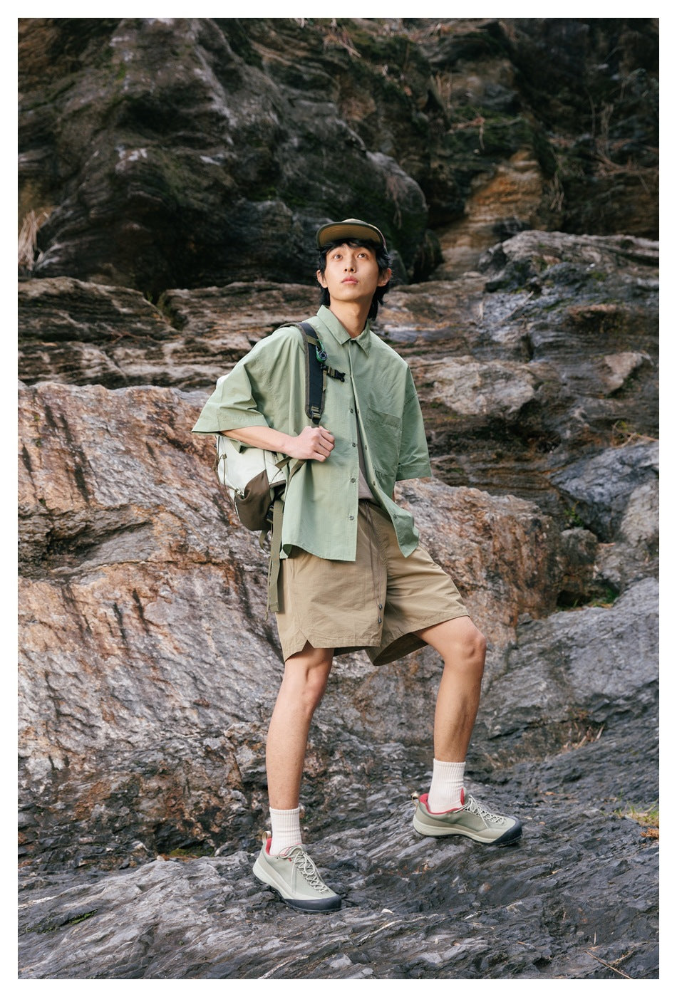Model wearing a pale sage oversized short-sleeve button-up shirt and tan shorts with a backpack on rocky terrain.