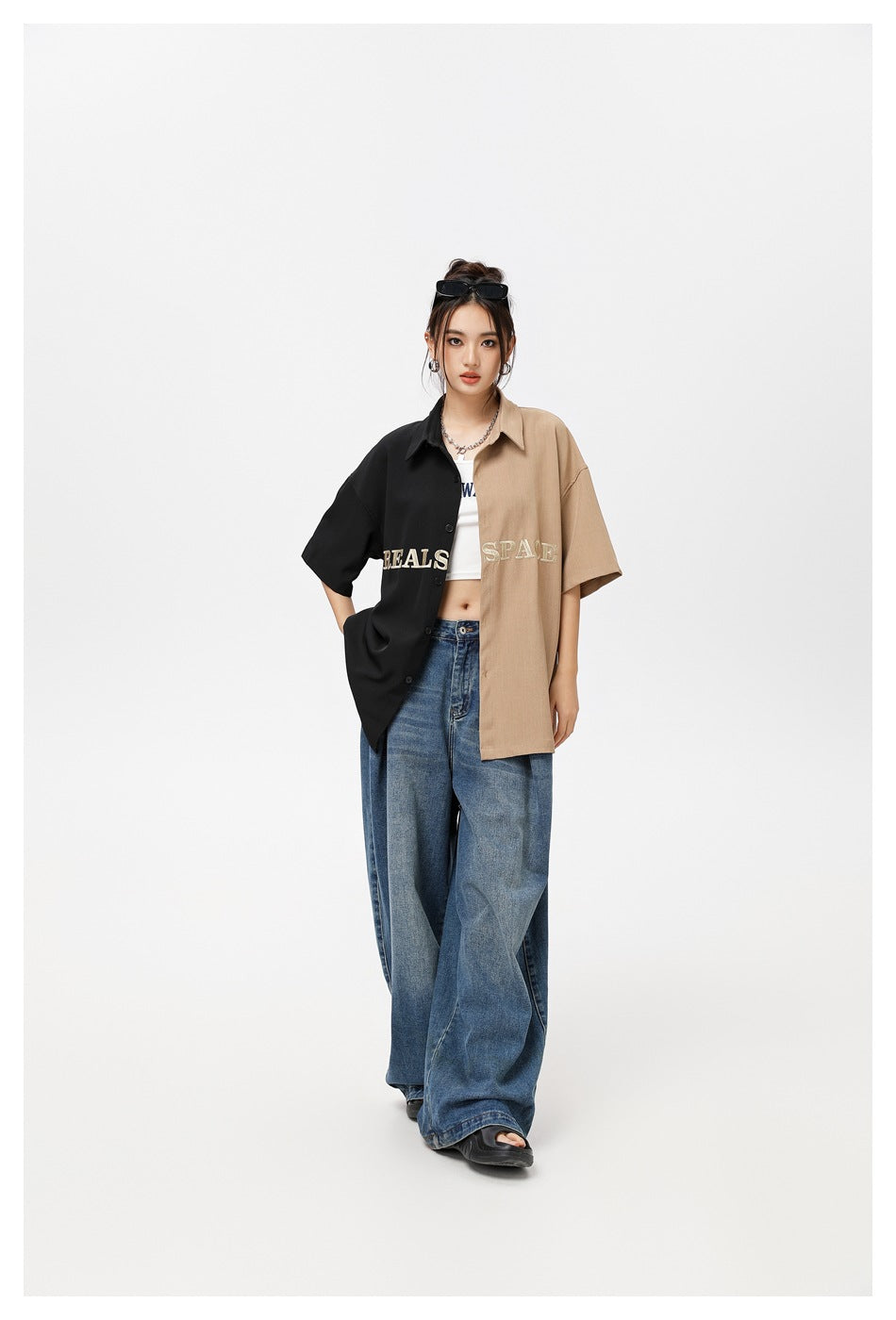 Model wearing an oversized black-and-tan embroidered shirt open over a white crop top and wide-leg blue jeans.