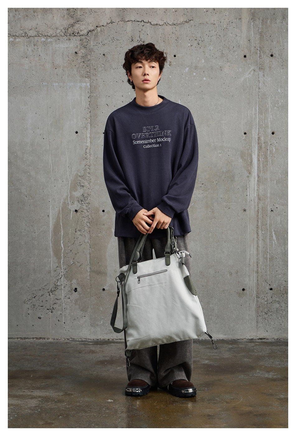 Man in a dark gray crew neck sweatshirt with chest lettering, wide pants and a light tote bag against concrete