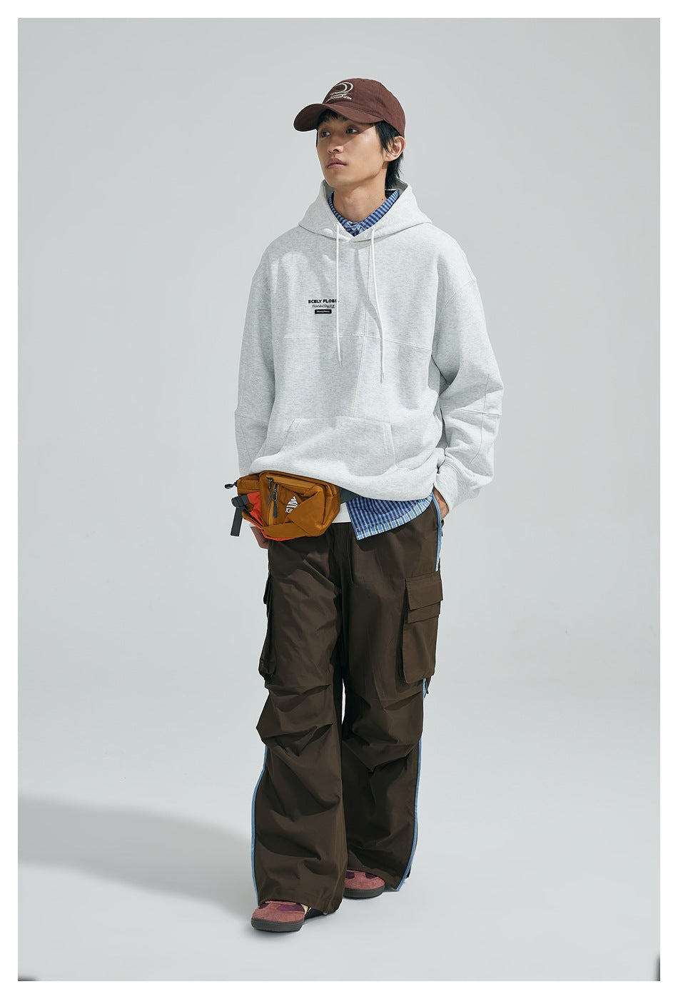 Model wearing a light gray hoodie over a striped shirt with brown cargo pants, orange waist bag and brown cap