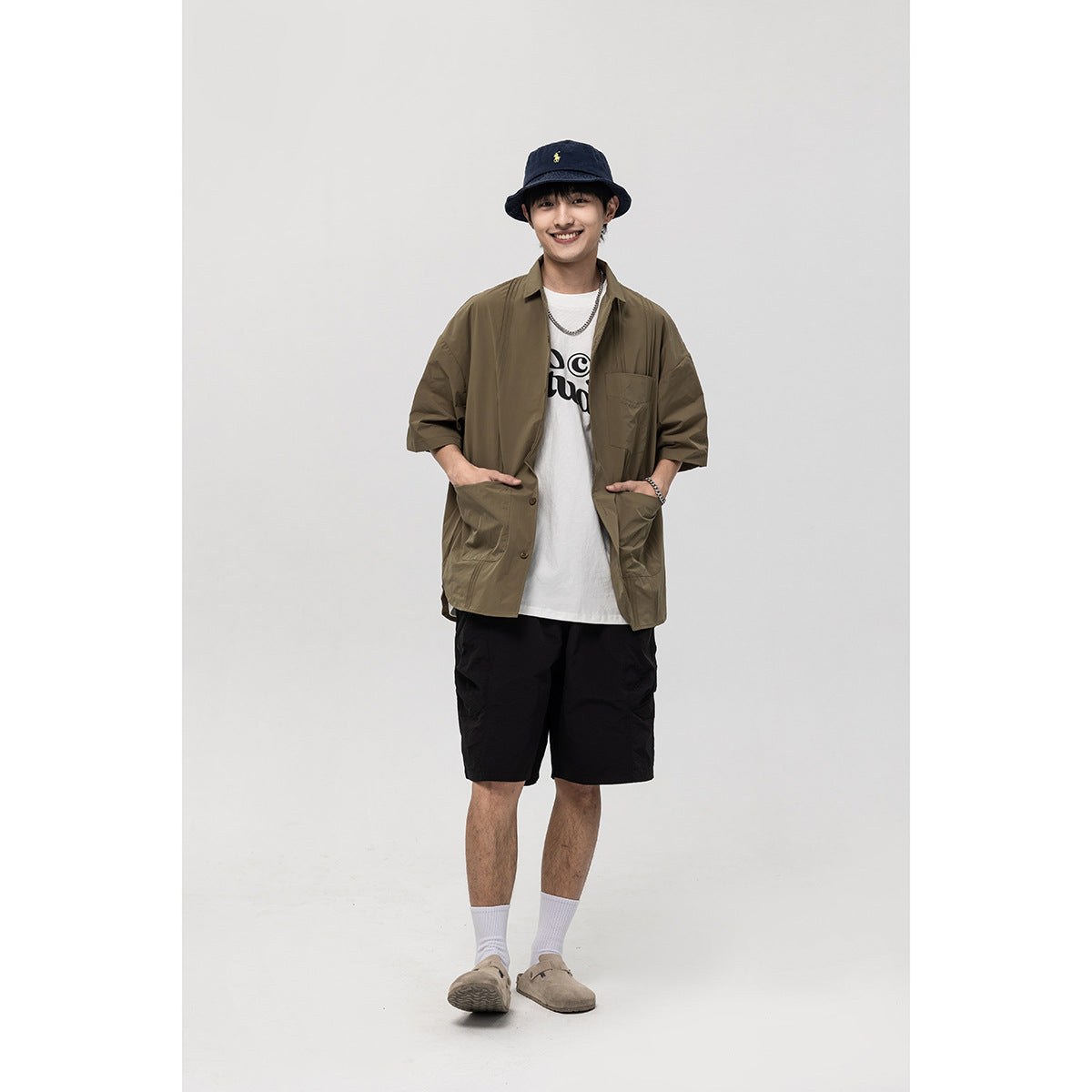 Model standing in an open olive short-sleeve shirt with chest and front pockets, layered over a white graphic T-shirt.