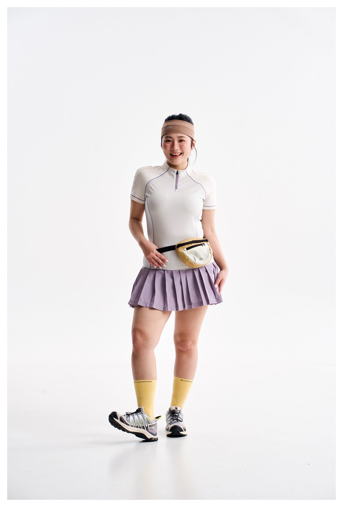 Model wearing a white short-sleeve zip top, pleated lilac skirt, yellow socks and a cream waist bag.