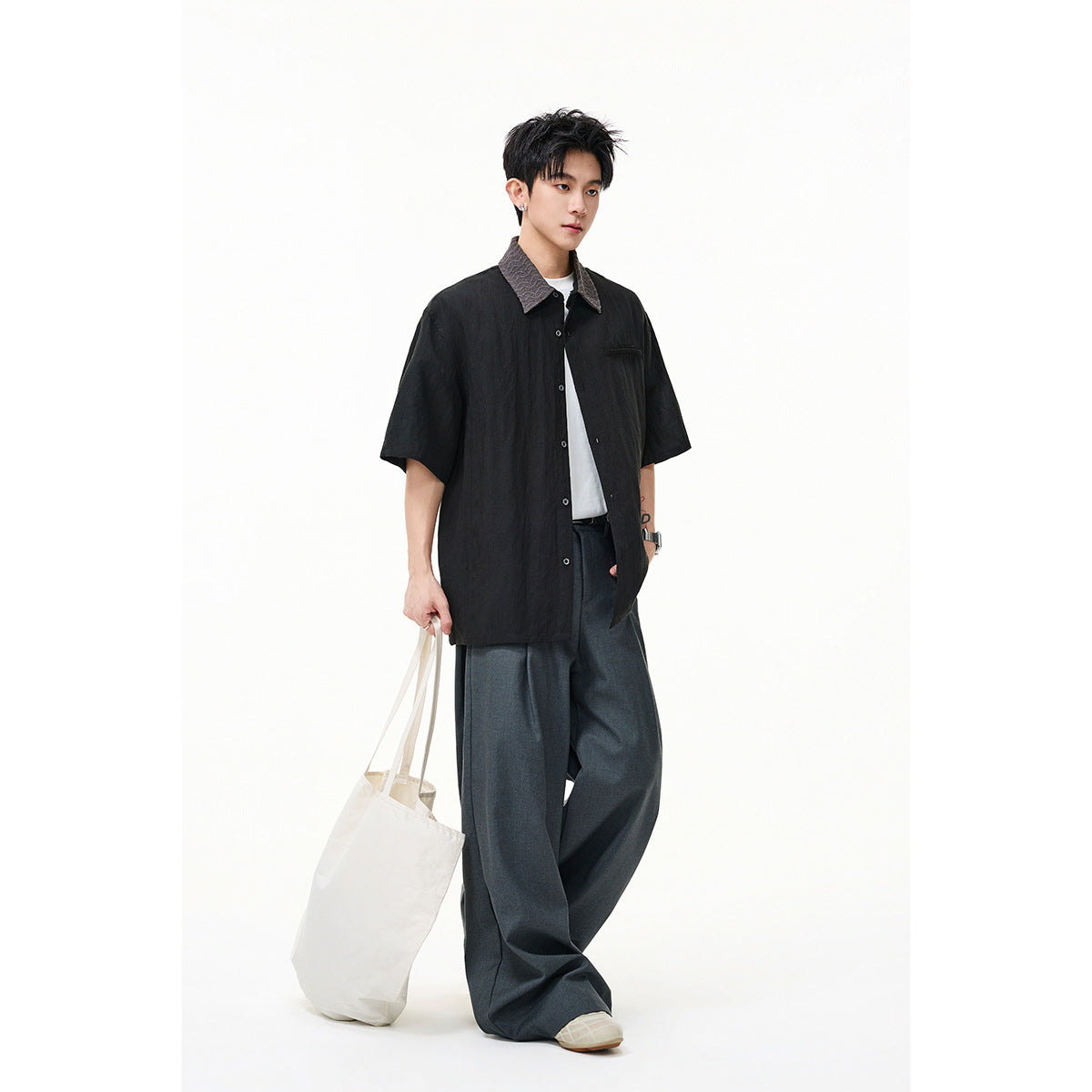 Model wearing a black short-sleeve button-up shirt with a textured gray collar, wide charcoal trousers, and a white tote.