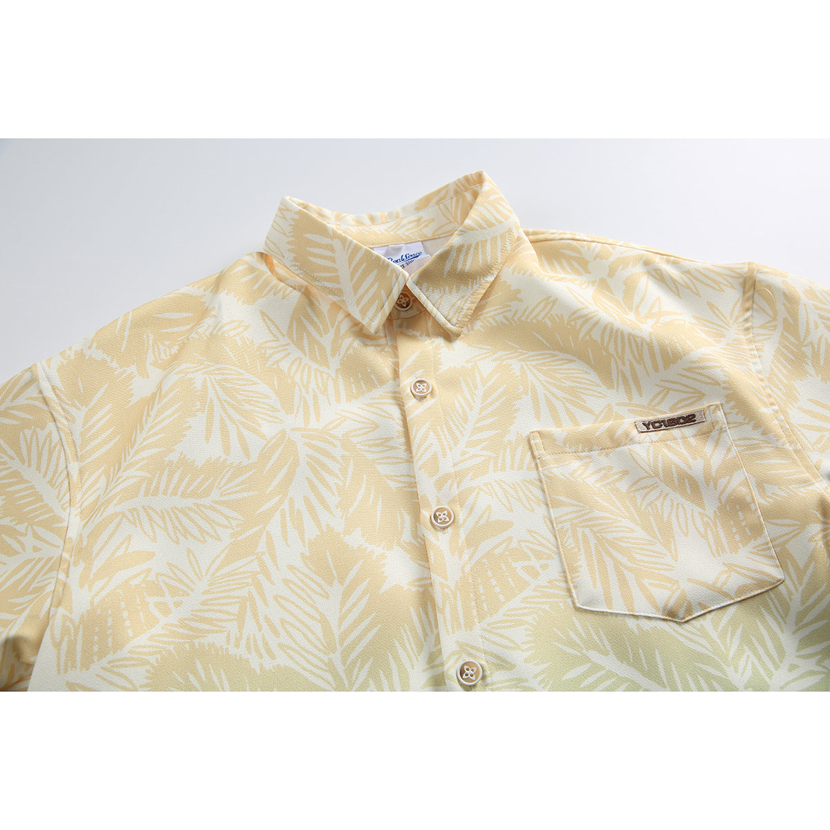 Close-up of a pale yellow tropical leaf print shirt, showing its collar, buttons, chest pocket, and small patch.
