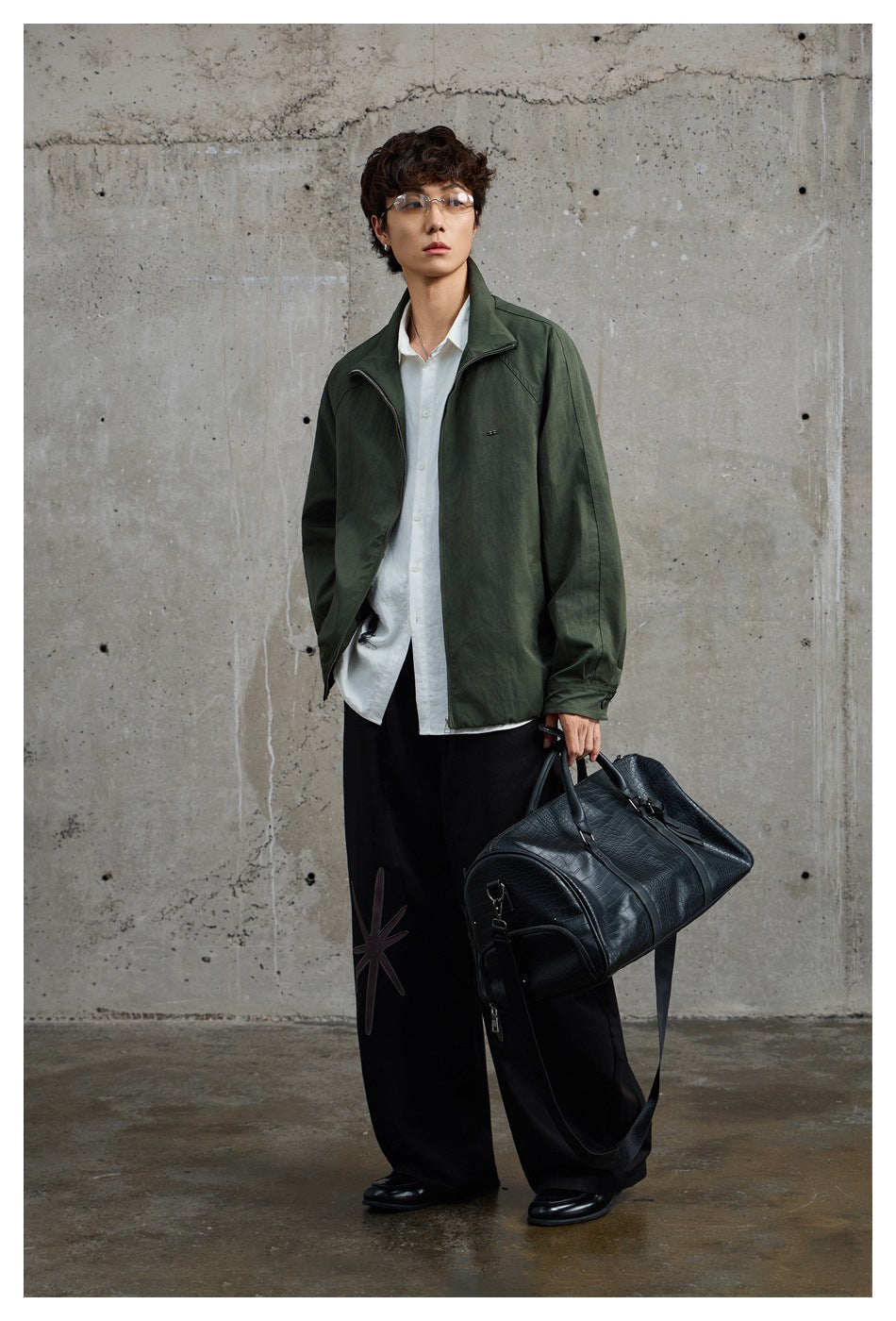 Man wearing an olive green zip jacket over a white shirt and wide black pants, holding a black duffel bag