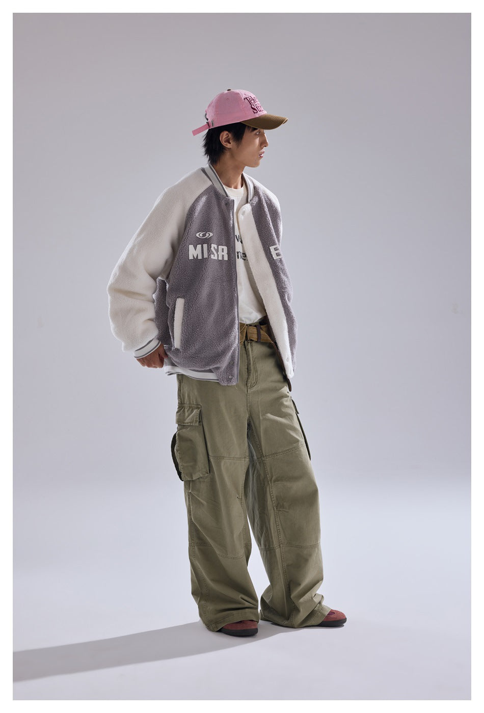 Person wearing the gray and cream baseball jacket open over a white tee with olive cargo pants and a pink cap