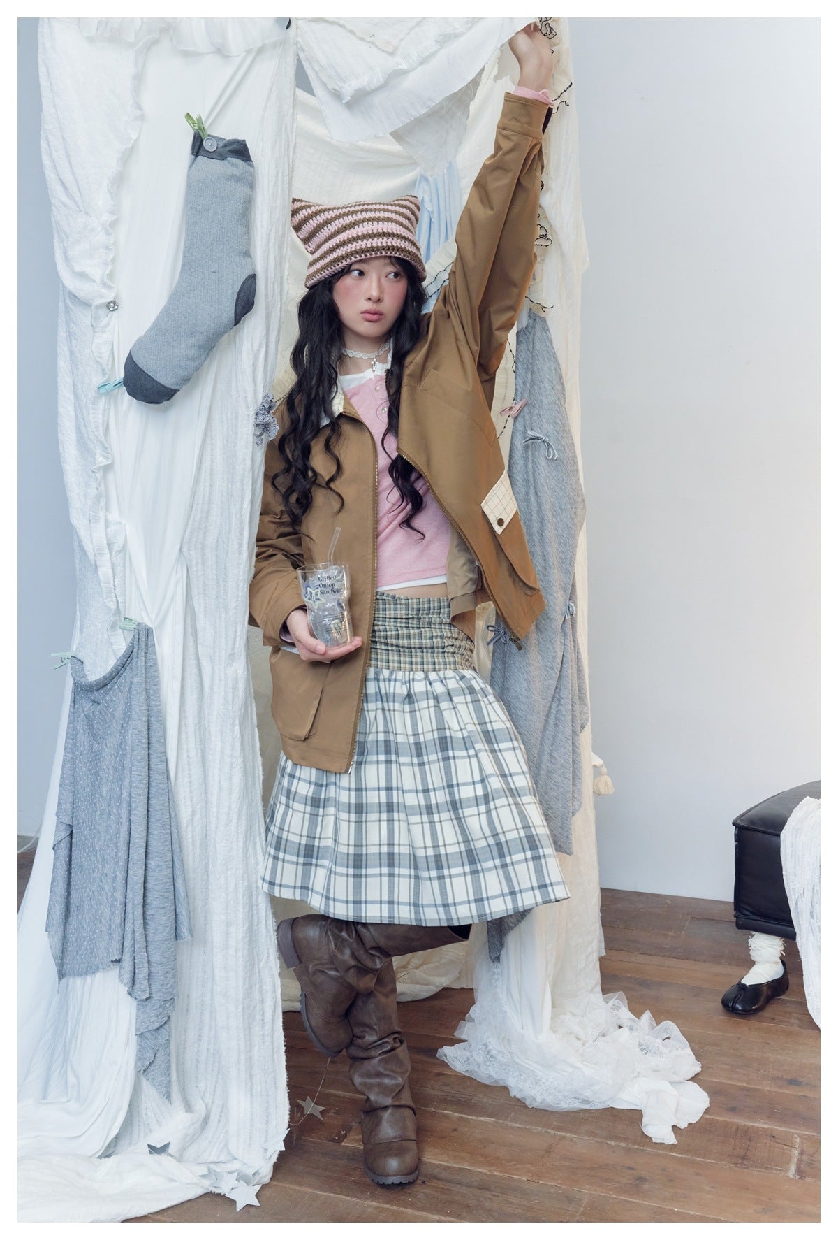 Model standing in a tan jacket with plaid details, pink top, and full plaid skirt, wearing a striped knit cat-ear hat.