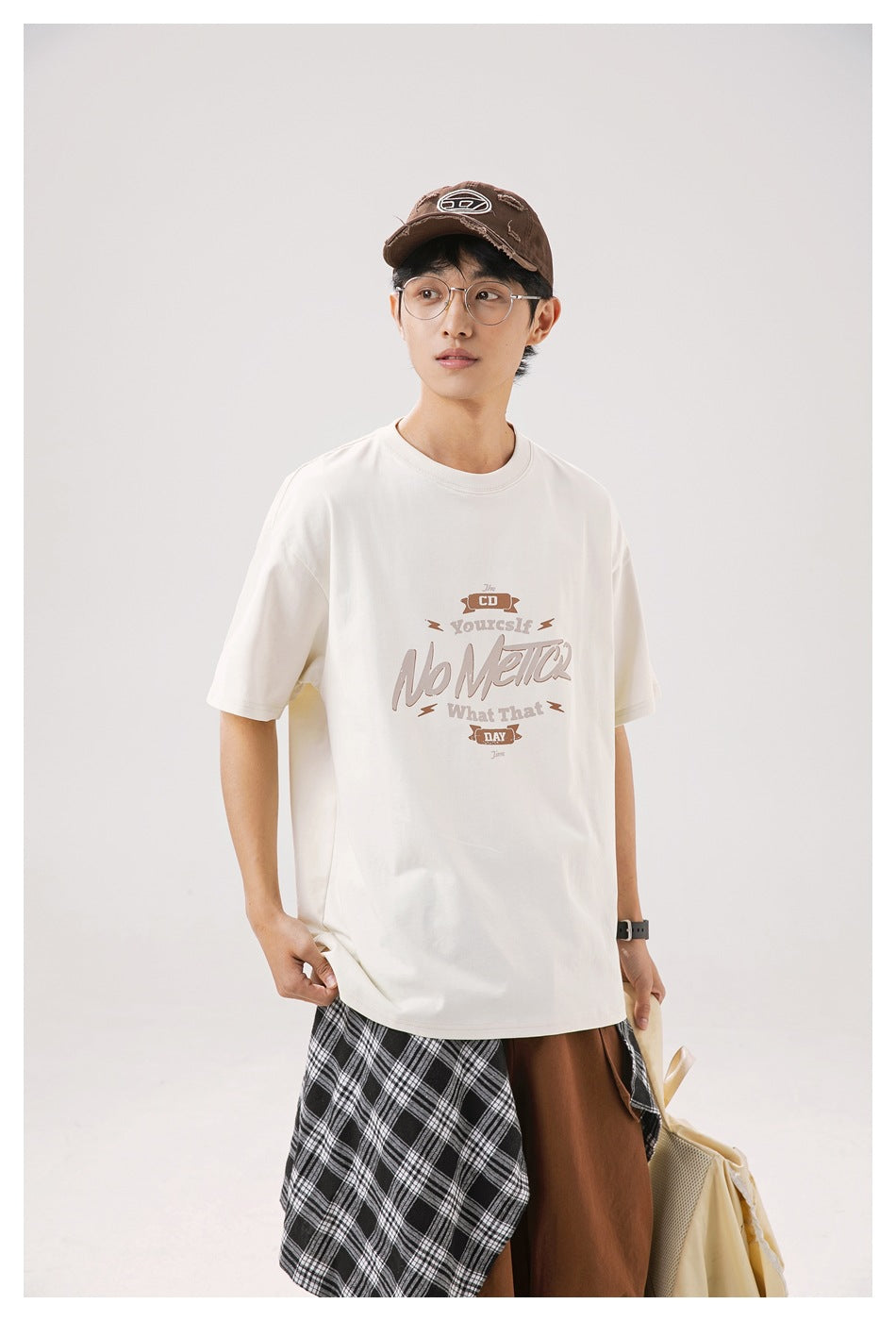 Model wearing an ivory oversized graphic T-shirt, shown from the front with brown trousers and plaid fabric.