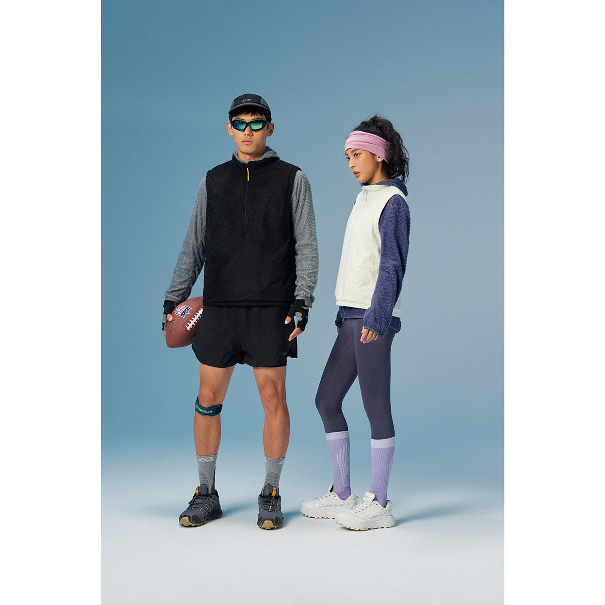 Two models wear layered athletic outfits, including a black pullover and shorts, and a cream vest with purple sleeves and