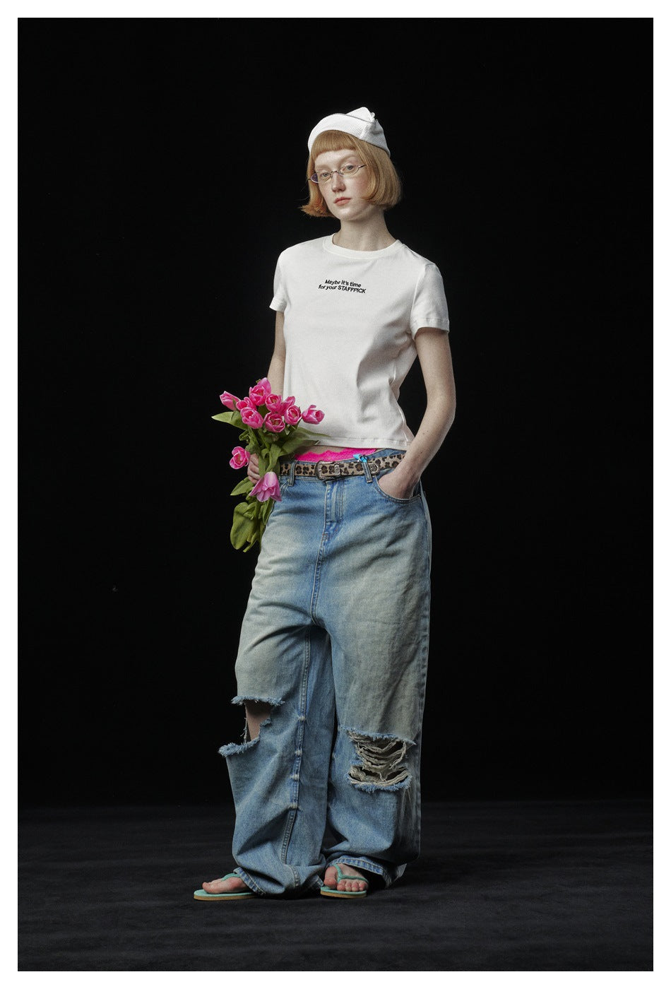 Model in a white slogan T-shirt and ripped wide-leg jeans, holding pink tulips against a black background.