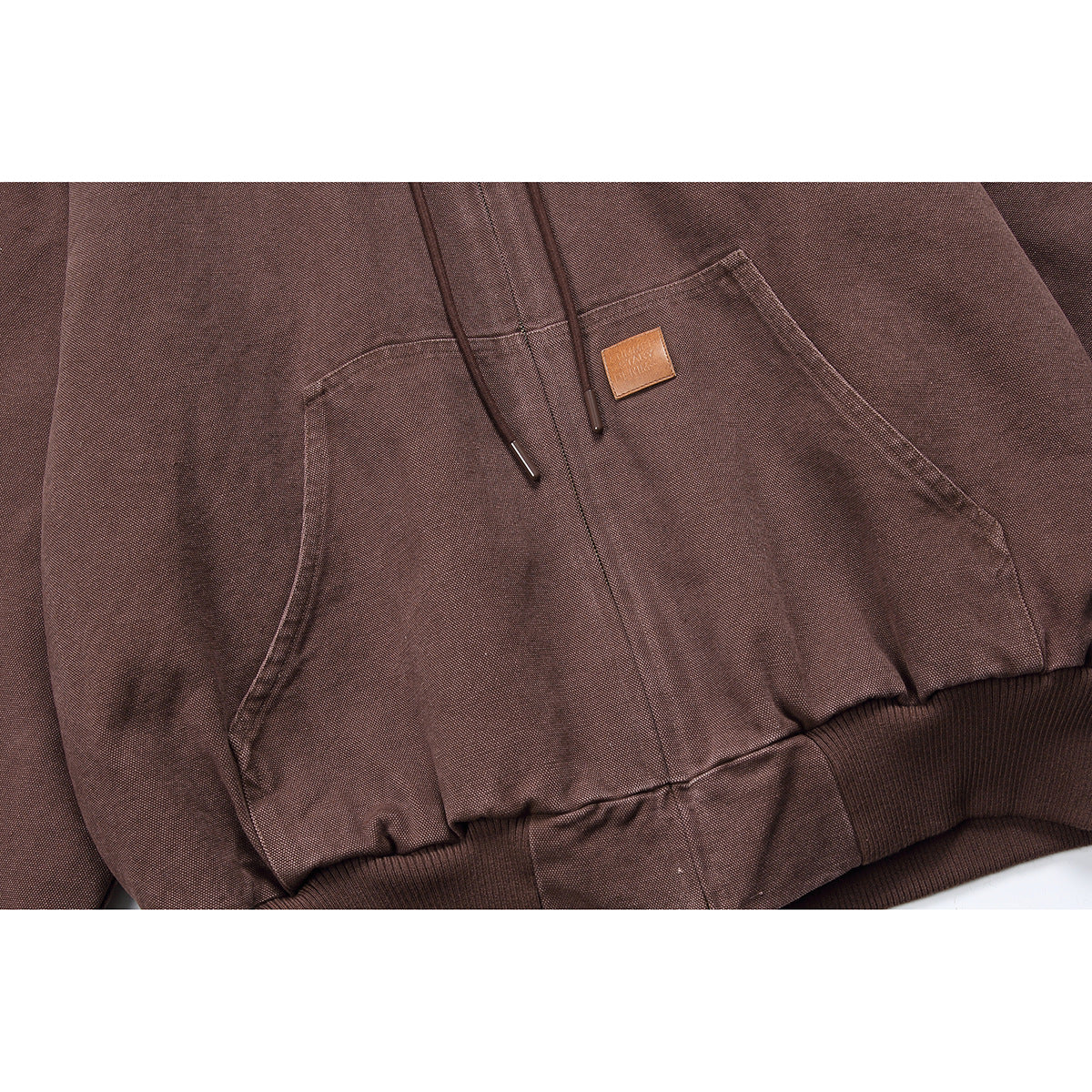 Close-up of a brown hooded jacket's drawstrings, kangaroo pocket, small patch, and ribbed hem.
