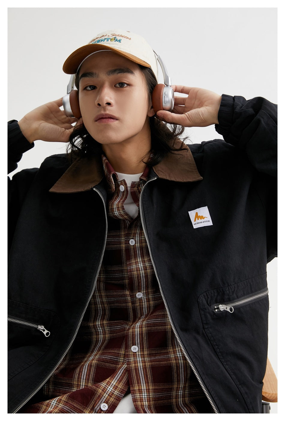 Model wearing a black zip-front jacket with a brown collar and zip pockets, layered over a brown plaid shirt.
