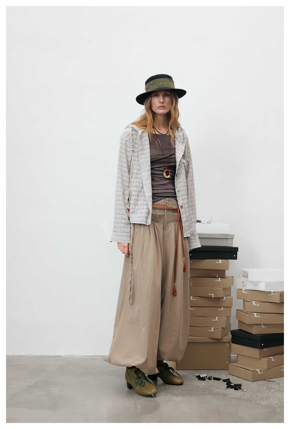 Model wearing the open gray plaid hooded shirt jacket over a brown top with wide beige trousers and a black hat