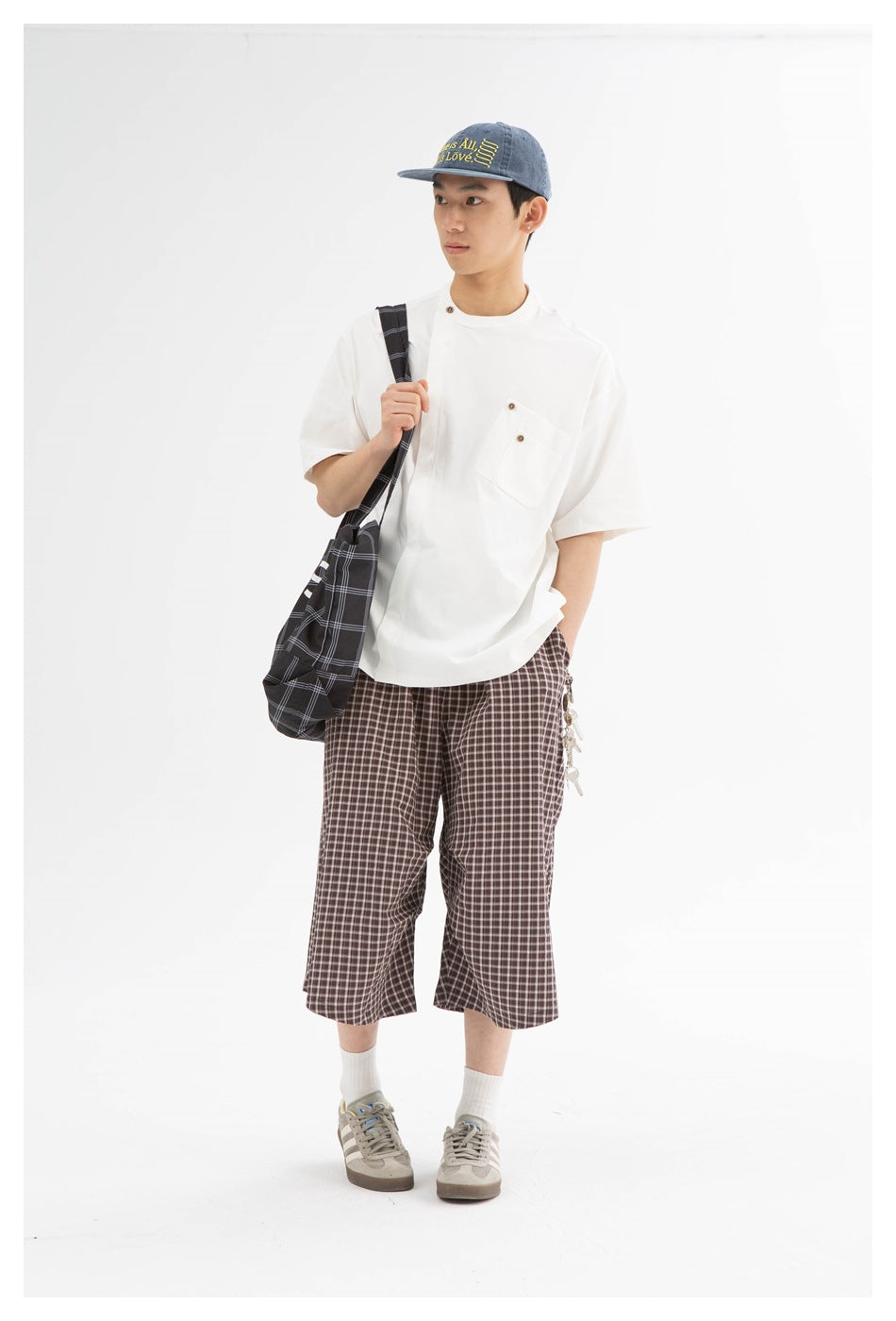 Model wearing a white short-sleeve shirt with a buttoned chest pocket and brown plaid shorts, carrying a dark plaid bag.