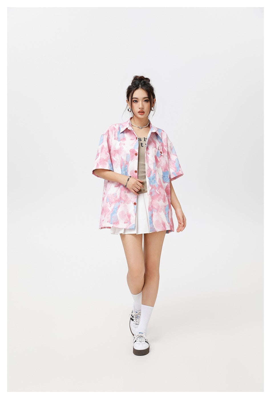 Model wearing a pink and light blue watercolor print short-sleeve shirt open over a tank top and white pleated skirt
