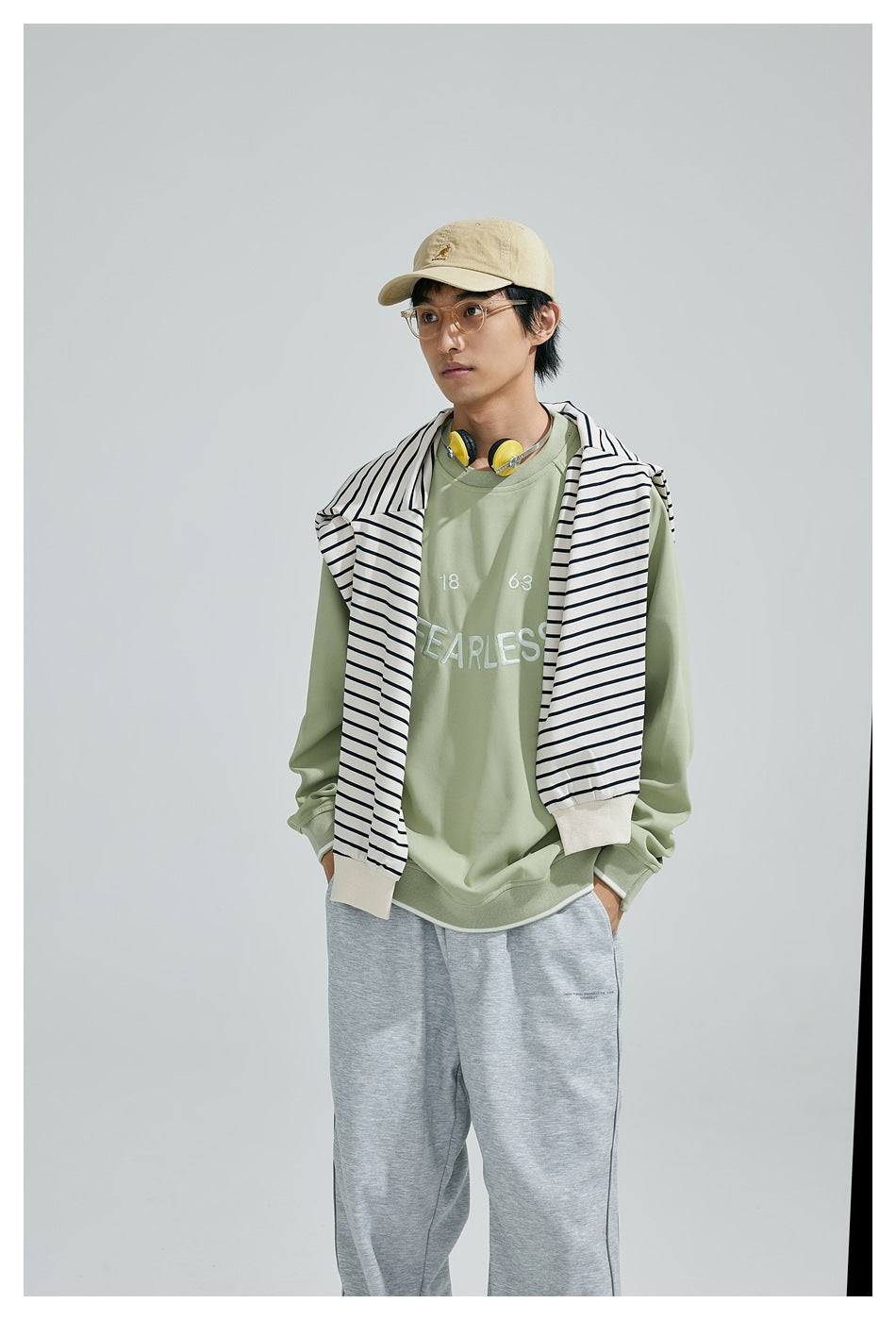 Model wearing a sage green FEARLESS sweatshirt with a striped top draped over the shoulders and gray pants.