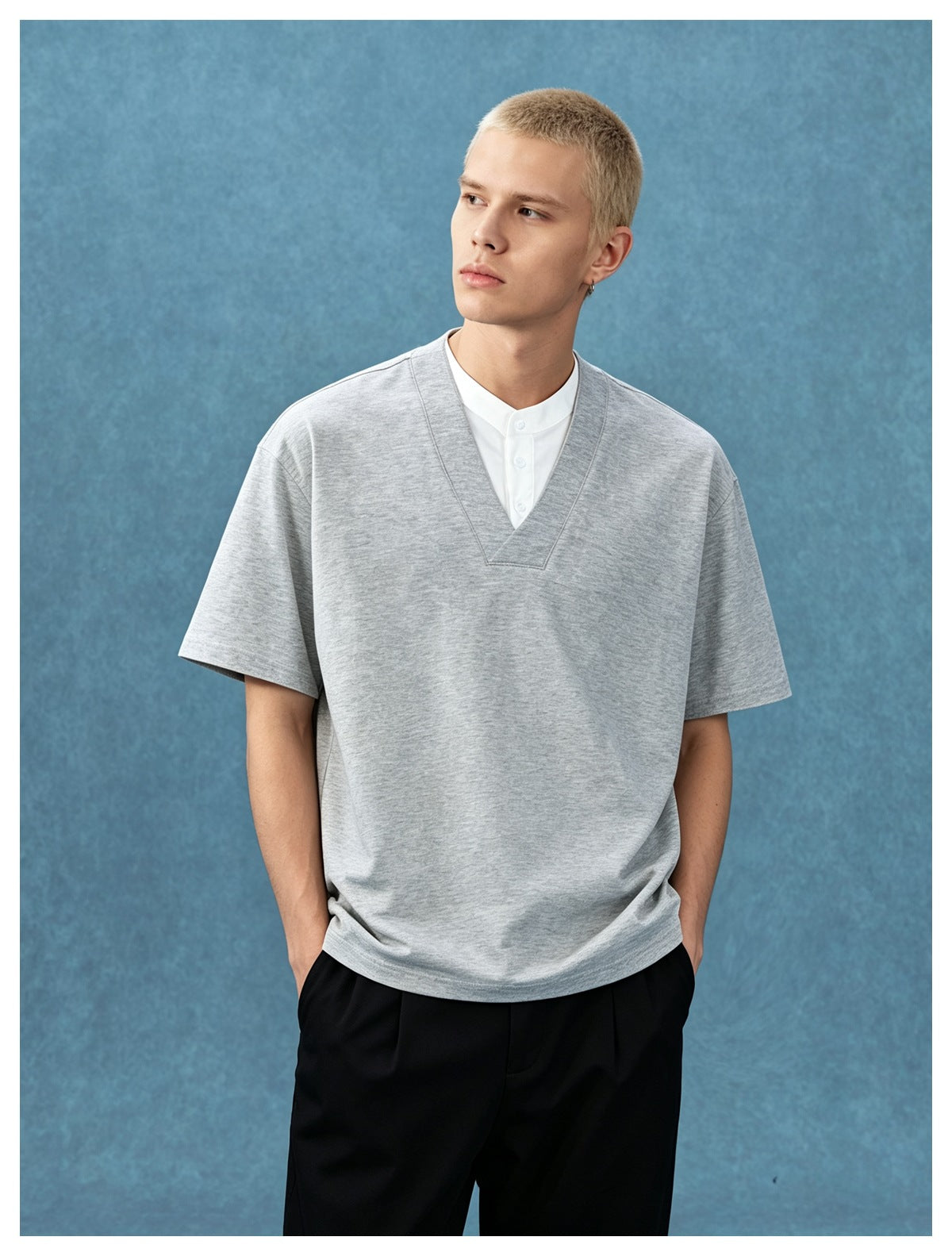 Man wearing a heather gray V-neck short-sleeve T-shirt over a white band-collar layer with black trousers