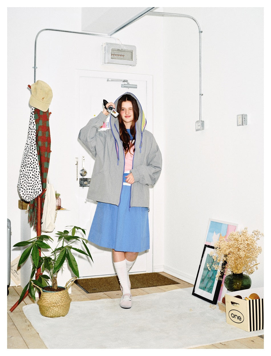 Person wearing the gray hooded jacket with the hood up over a pink top and blue skirt in a white room