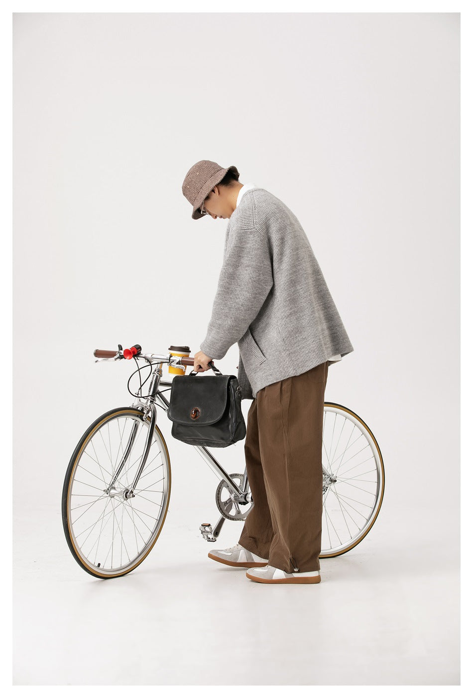 Side view of a model wearing a light gray textured cardigan and brown trousers beside a bicycle.
