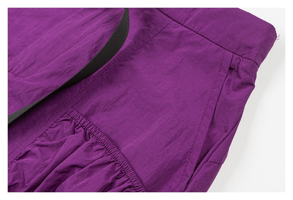 Close-up of purple pants with a black strap, stitched panel seam, and gathered pocket edge.