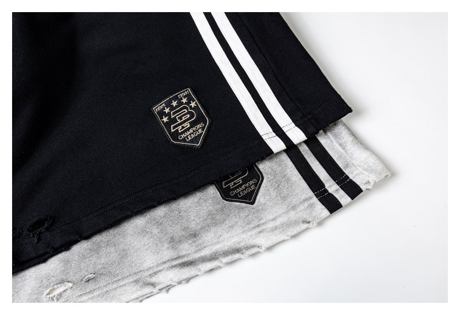 Close-up of black and gray athletic shorts hems with twin side stripes, embroidered shield patches, and distressed edges.