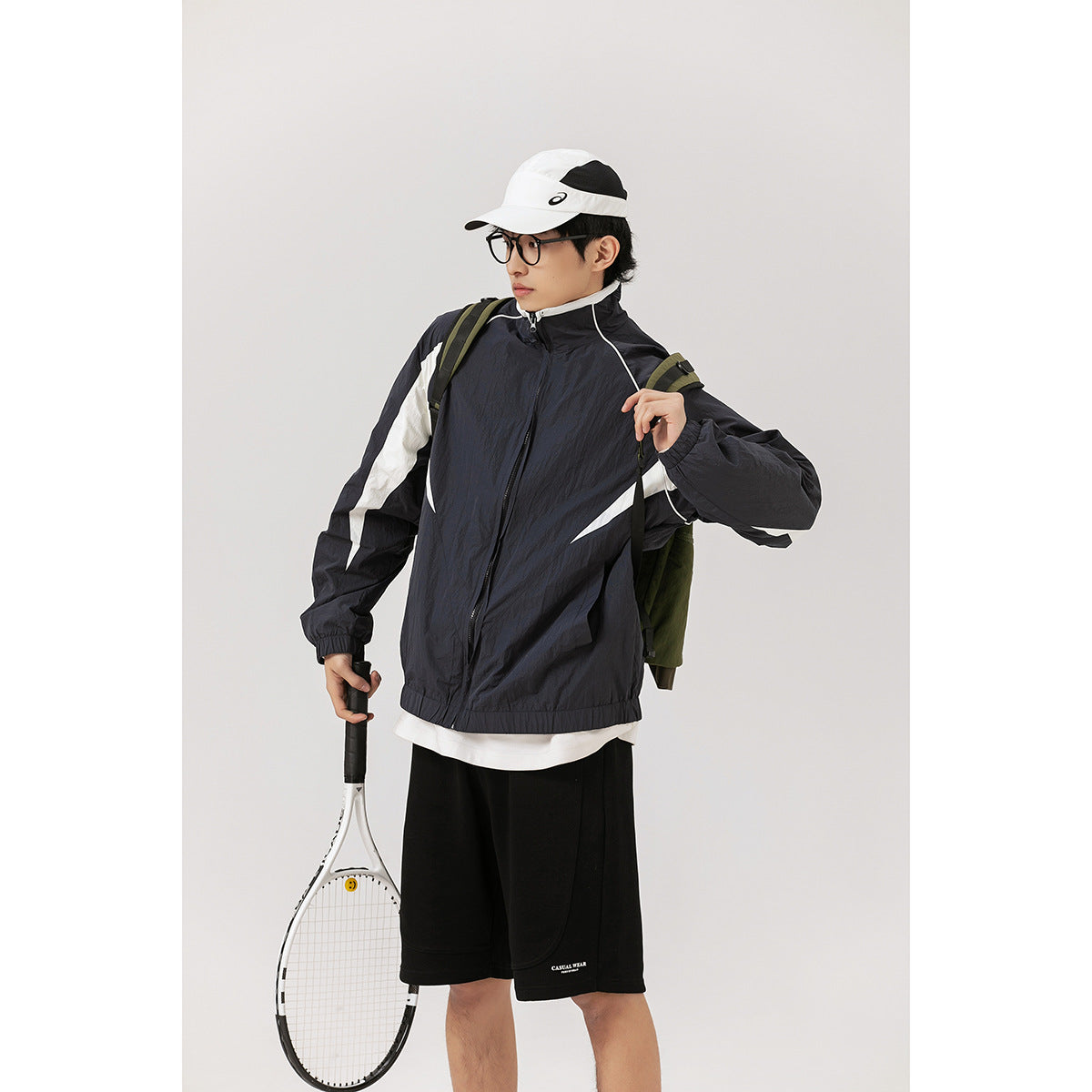 Model wearing a black zip jacket with white sporty panels, black shorts and a white cap, holding a tennis racket.