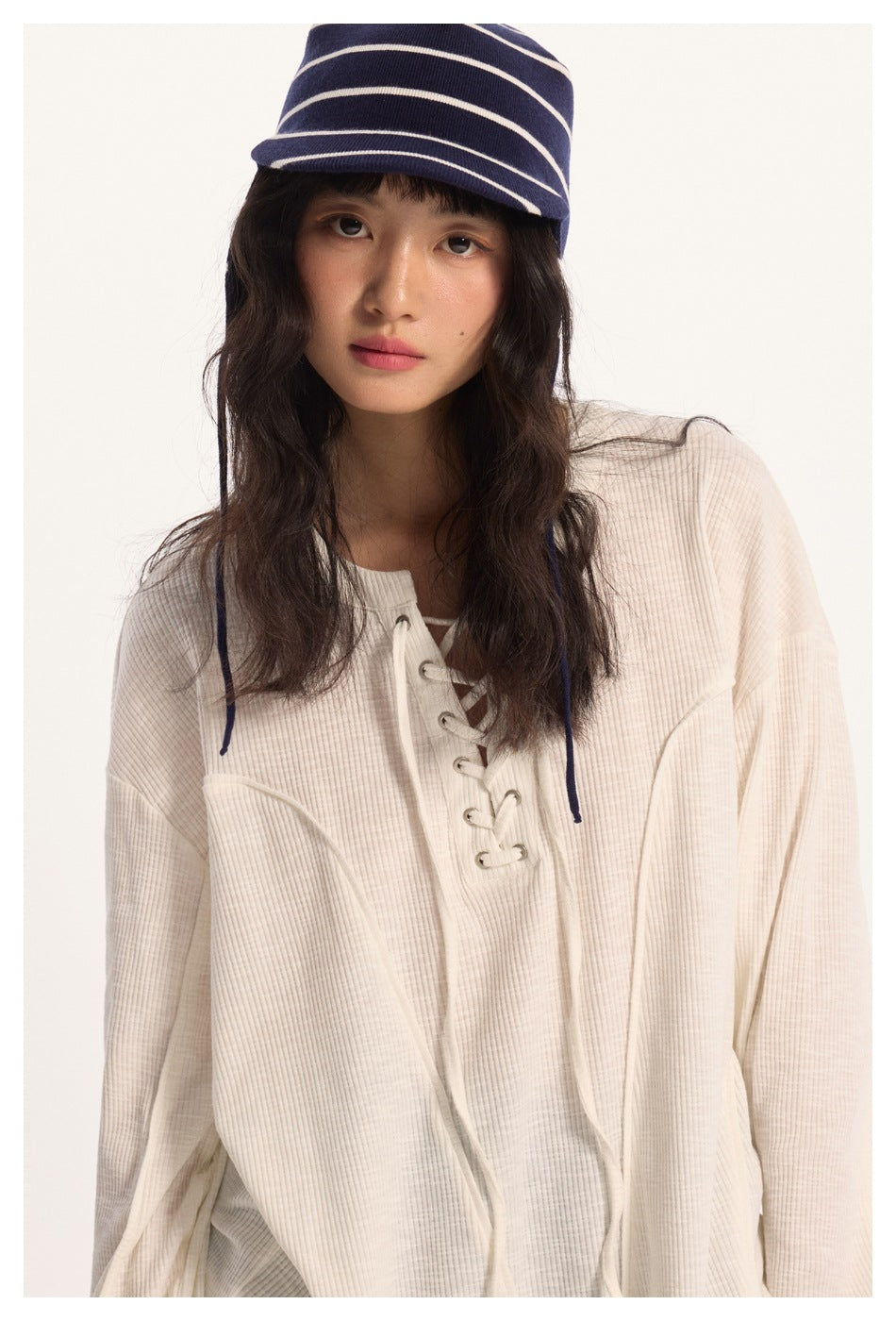 Model wearing a cream ribbed long-sleeve top with a lace-up front and a navy striped hat.