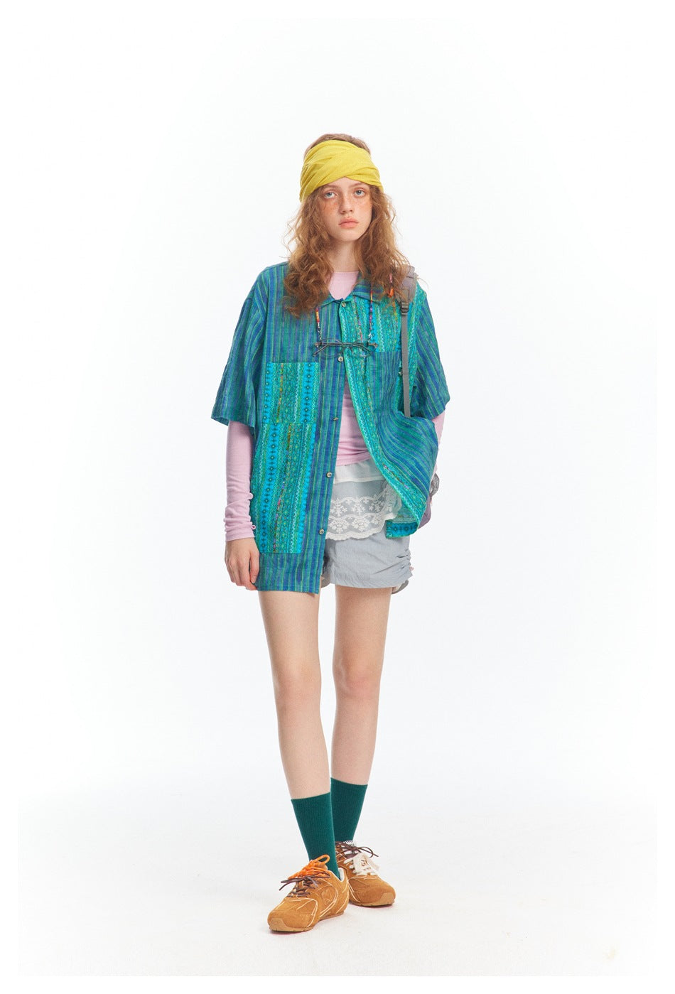 Full-length model wearing a blue-green patterned short-sleeve shirt, pink sleeves, gray shorts, and a yellow headband.