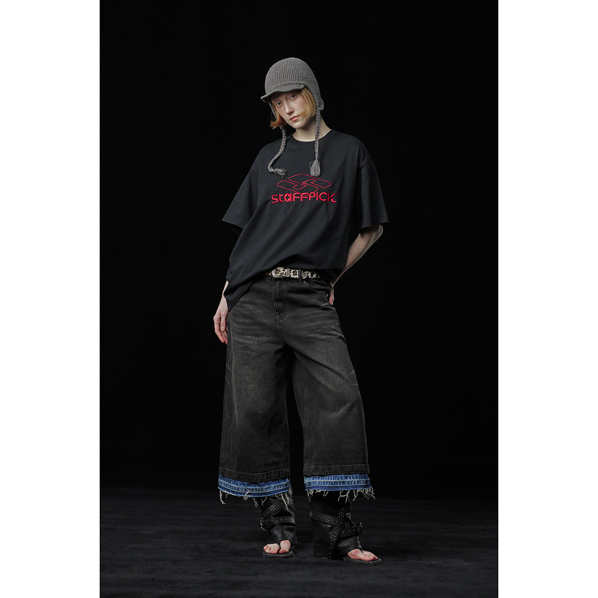 Model wearing an oversized black T-shirt with red chest lettering, wide dark jeans, and a gray knit cap.