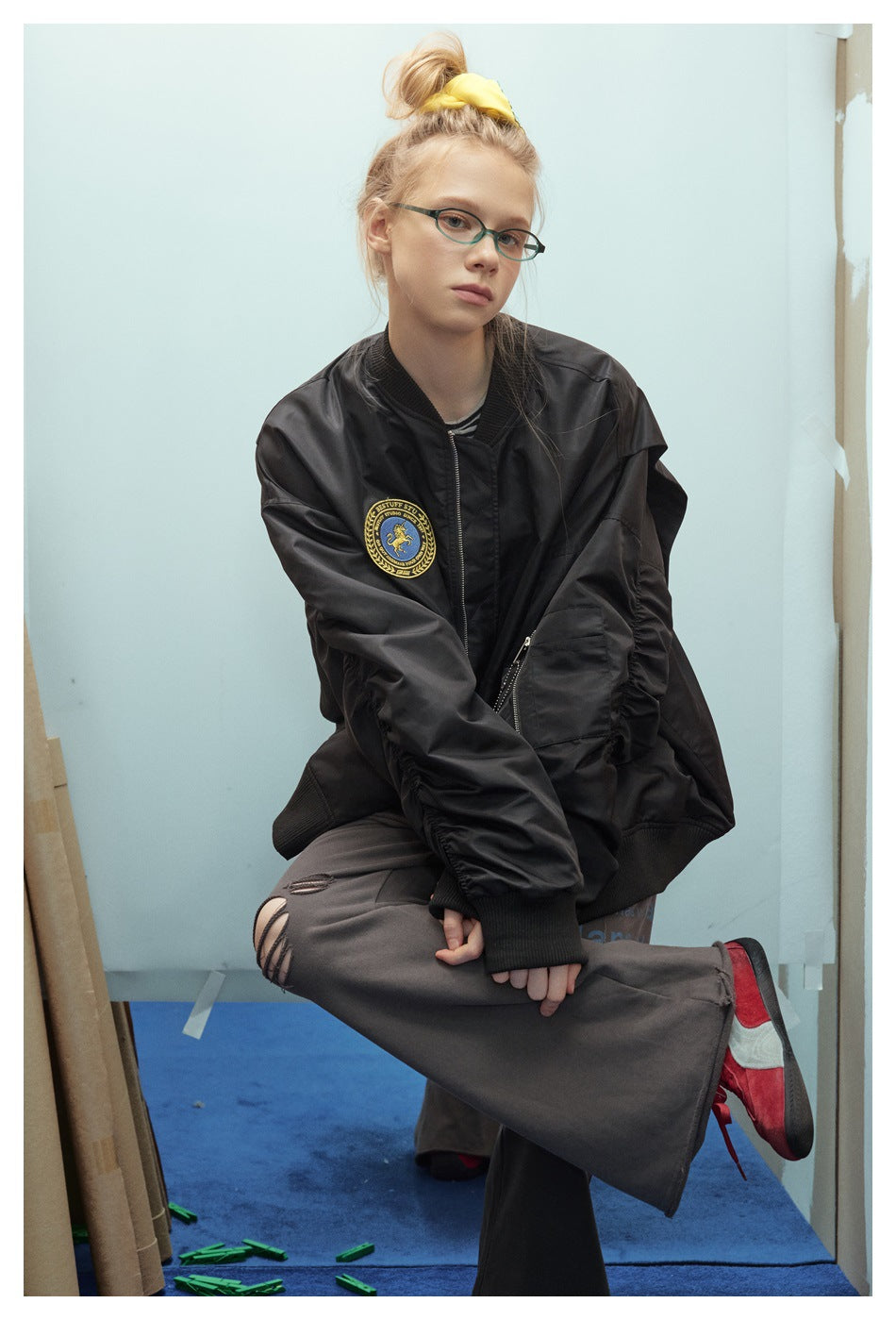 Model seated in an oversized black bomber jacket with a gold-and-blue chest patch, ribbed cuffs, and zip pockets.