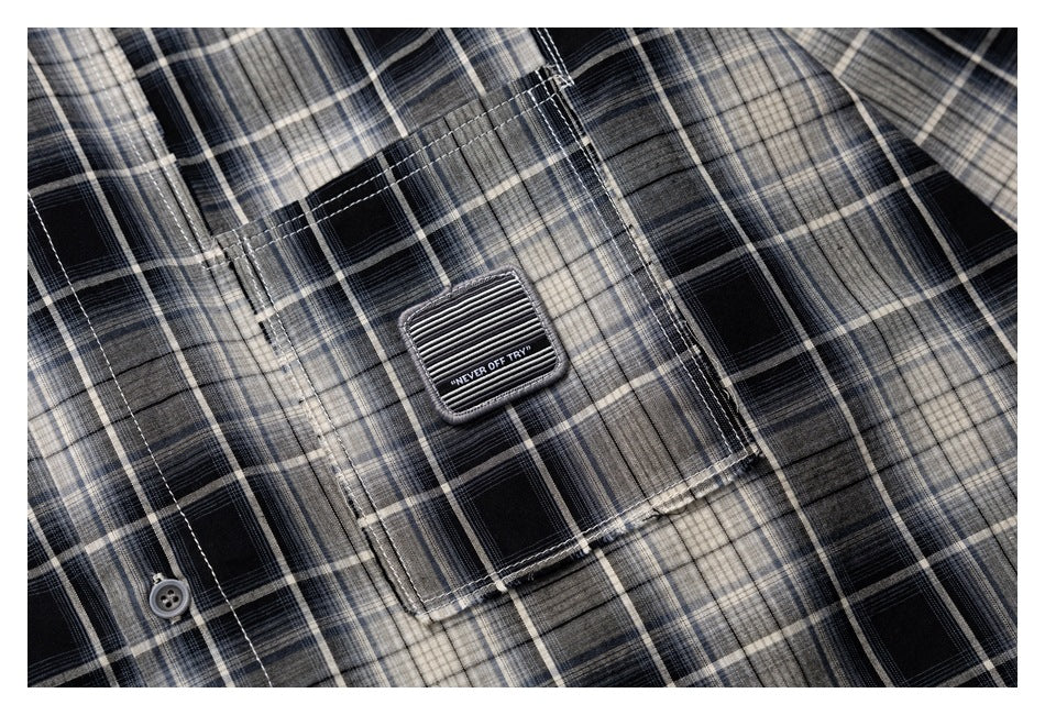 Close-up of a black, gray, and cream plaid shirt pocket with a stitched patch and button.