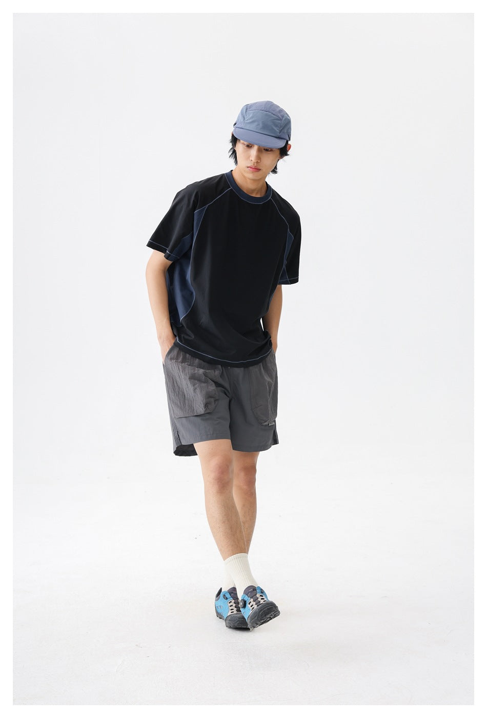 A man models a black and navy color-block T-shirt and gray utility shorts, standing at an angle against a white background.