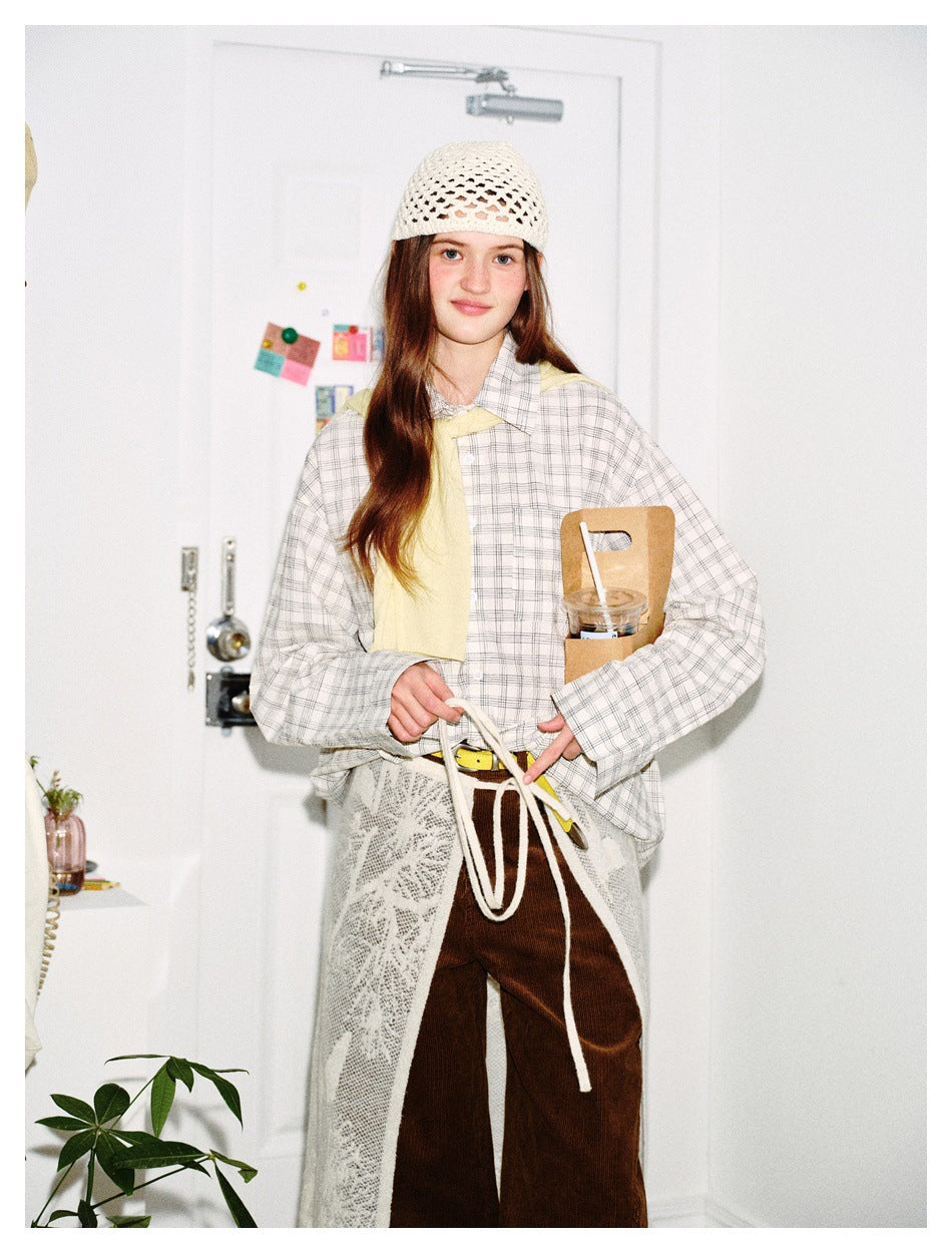 Front view of a model in a light plaid shirt, brown pants, white lace apron and crochet cap, holding an apron tie.