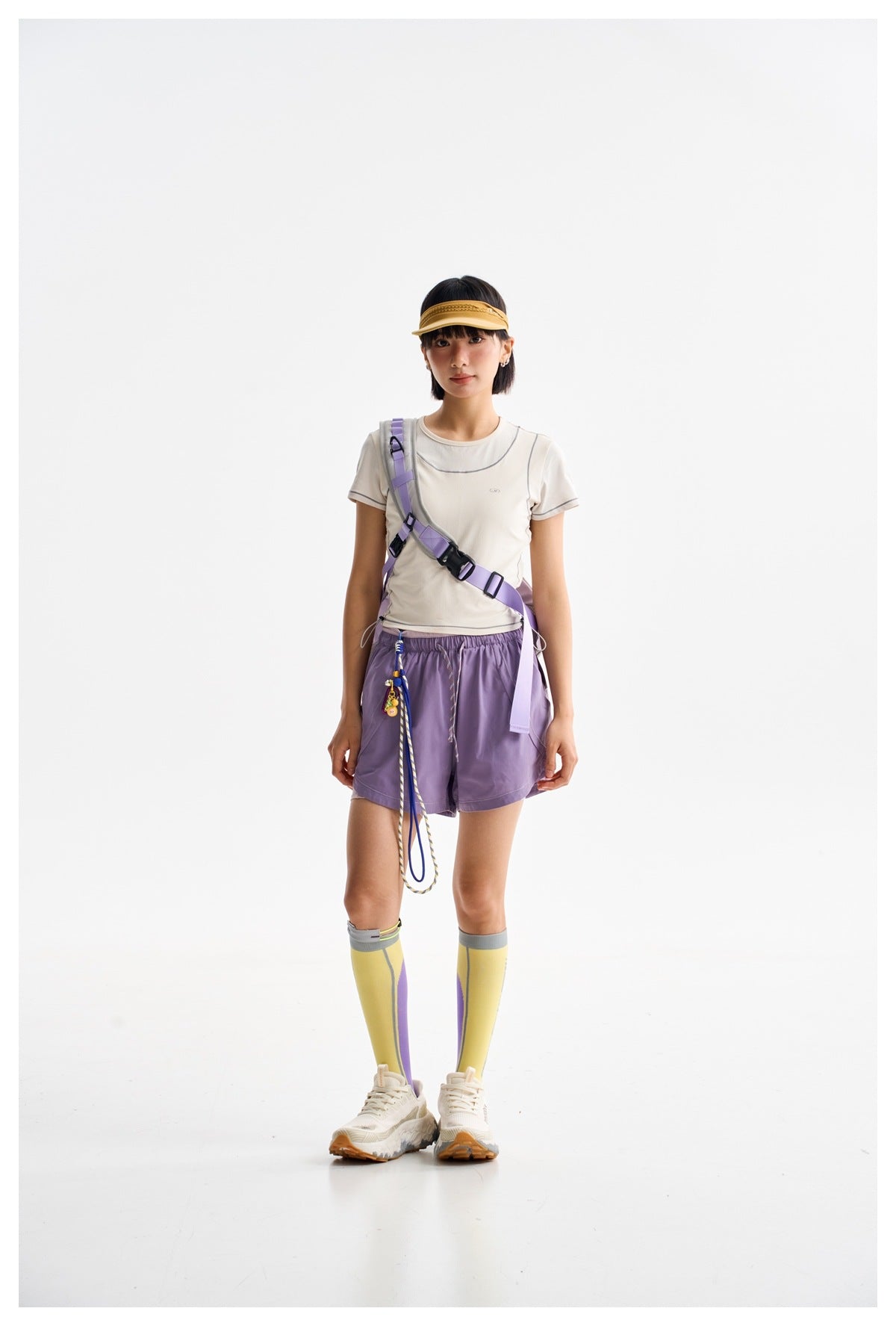 Model wearing a cream athletic T-shirt, purple shorts, a lilac crossbody harness, yellow visor, and color-block knee socks.