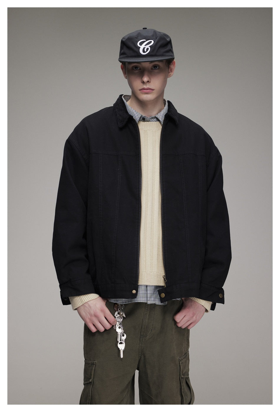 Model wearing a black zip-front jacket over a cream sweater and checked shirt, with olive cargo pants and a black cap.