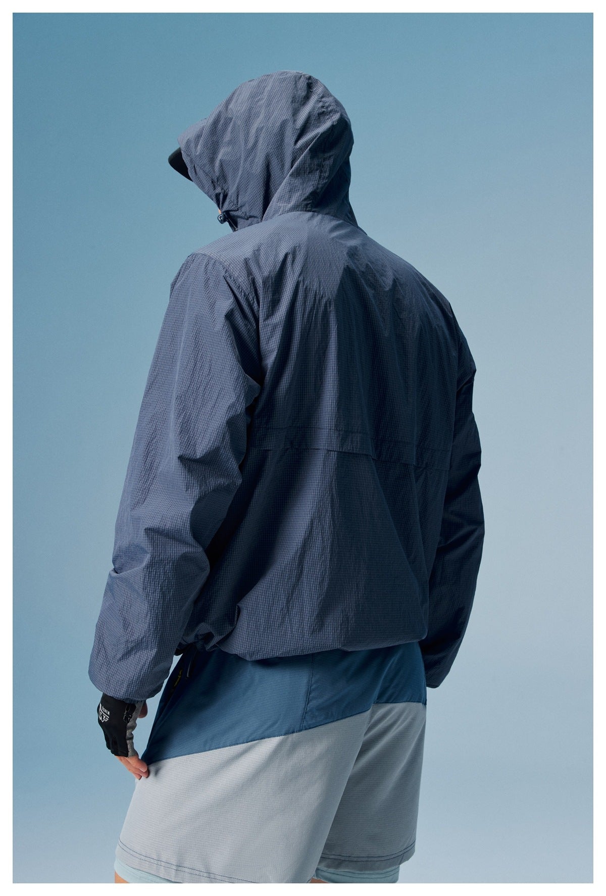 Back view of a model wearing a slate-blue hooded anorak with a fine grid pattern and gray shorts.