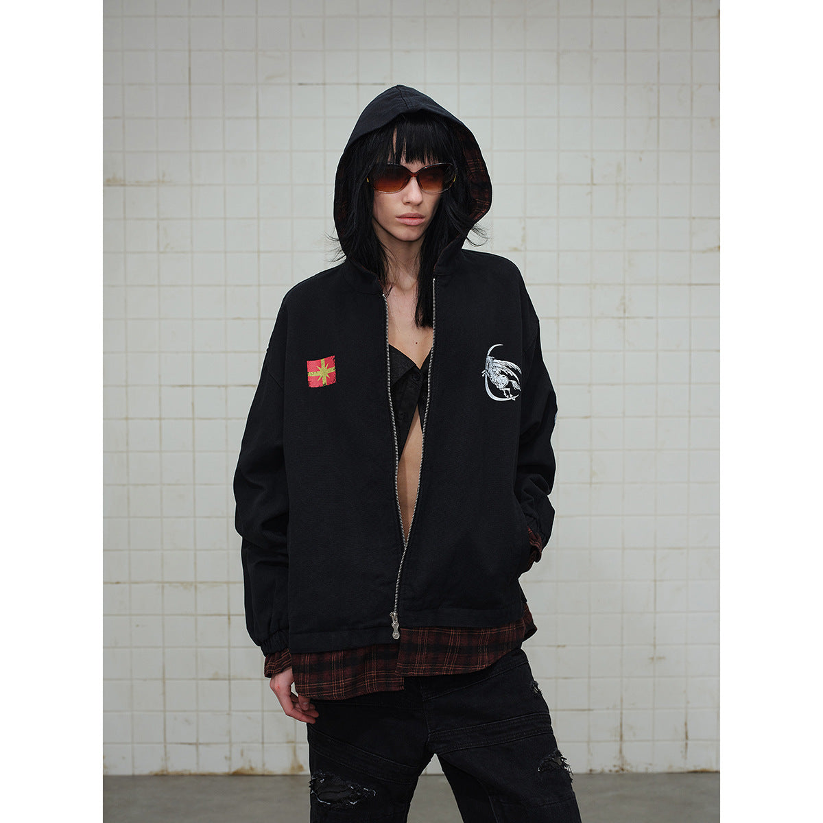 Front view of a model wearing a black hooded zip jacket with chest graphics and a red plaid layer underneath.