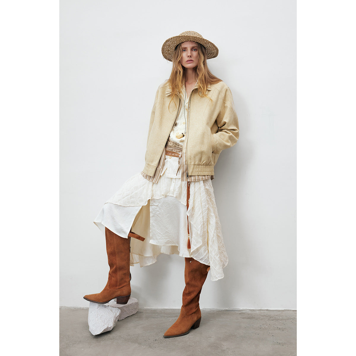 Full-length model wearing a pale beige zip jacket over a layered white skirt, with brown boots and a straw hat.