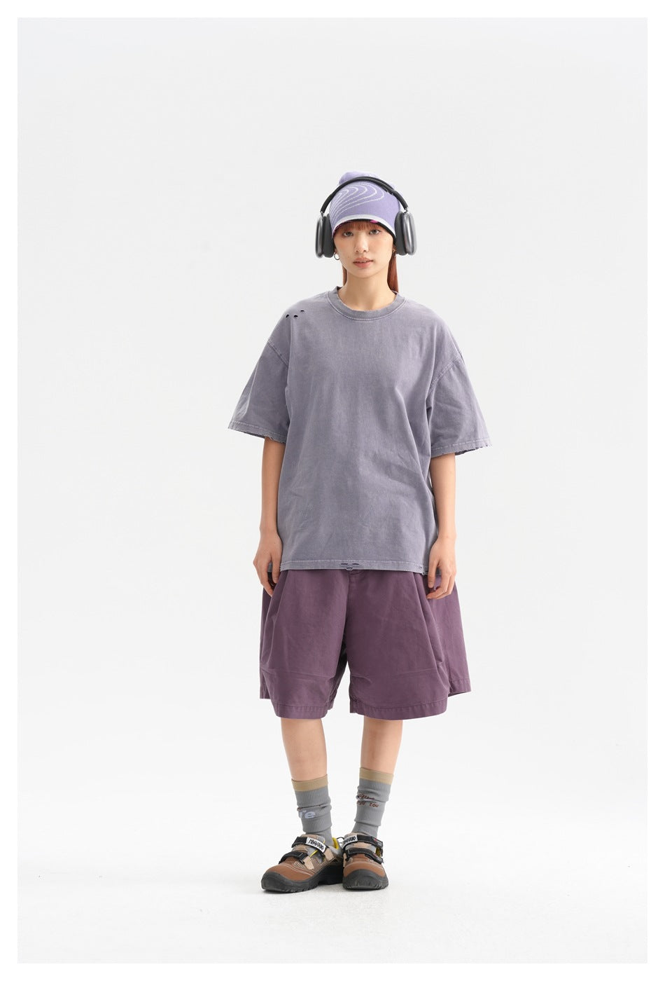 Model wearing an oversized faded lavender T-shirt and plum shorts, with a patterned cap and over-ear headphones.