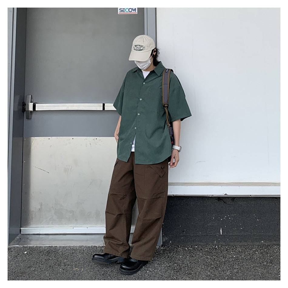 Person wearing a green short-sleeve shirt open over a white tee with brown cargo pants and a cap
